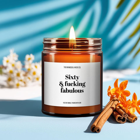 Close-up of a lit amber glass soy candle on a white surface with soft shadows. The label reads “Sixty & Fucking Fabulous” in bold black font, designed by The Whimsical Wicks Co. A bouquet of tulips in the background adds a cozy and elegant atmosphere, perfect for a sassy 60th birthday gift with attitude.