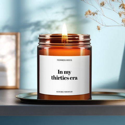 Amber glass jar candle with the label “In My Thirties Era” lit beside cinnamon sticks and orange blossoms, styled on a beachy pastel background.
