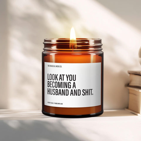 Close-up of a 9oz amber glass jar candle with black metal lid, white label reading “Look at You Becoming a Husband and Shit” in bold black text, styled on a neutral background with warm lighting, perfect for funny groom gifting.