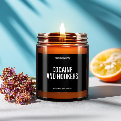Amber glass jar candle with a pink label that reads “Cocaine and Hookers” in bold text, lit and placed on a smooth stone with dried lavender nearby, styled against a soft aqua background. Playful, edgy, and perfect for adult humor gifts or novelty candle lovers.