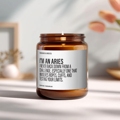 Amber glass candle with the label "I’m an Aries – I never back down from a challenge, especially one that involves ropes, cuffs, and testing your limits" styled on a sunlit shelf with soft background blur and cozy accents.