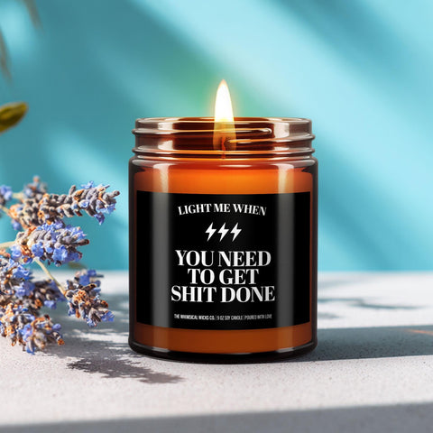 Amber jar candle with label that reads “Light when you need to get sh*t done,” lit and styled with lavender and citrus on a bright blue background.