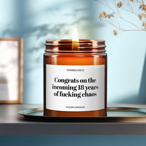 Amber glass candle labeled “Congrats on the Incoming 18 Years of Fucking Chaos,” styled with dried florals, citrus slices, and parenting books for a cheeky baby shower vibe.