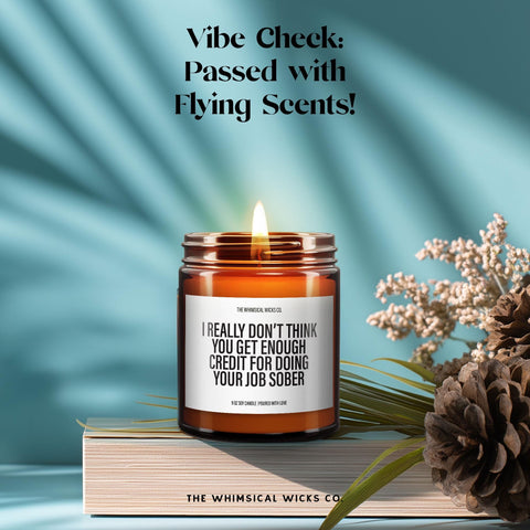 Humorous amber glass candle on a hardcover book beside pinecones and dried florals. Label reads “I really don’t think you get enough credit for doing your job sober.” Above the candle is the phrase: “Vibe Check: Passed with Flying Scents!” Playful and cheeky office gift.