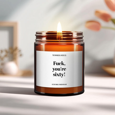 A lit amber glass candle with the label “Fuck, you're sixty!” styled with summery flowers and cinnamon sticks, perfect for milestone birthday celebrations and gag gifts.