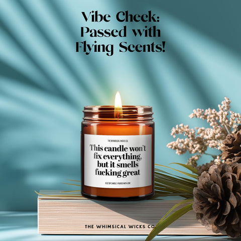 Amber glass jar candle labeled “This Candle Won’t Fix Everything But It Smells Fucking Great” placed on an open book with sprigs of lavender, styled for a cozy yet cheeky vibe, ideal for friends or coworkers who appreciate humor with their decor.