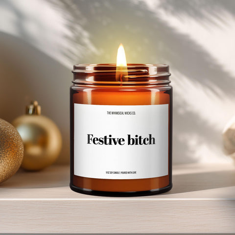 Close-up of a 9oz amber glass jar candle with black metal lid, white label reading “Festive Bitch” in bold black text, styled on a neutral background with warm lighting, perfect for cheeky and humorous holiday gifting.