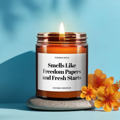 Close-up of a 9oz amber glass jar candle with black metal lid, white label reading “Smells Like Freedom Papers and Fresh Starts” in bold black text, styled on a neutral background with warm lighting, perfect for funny divorce gifting.
