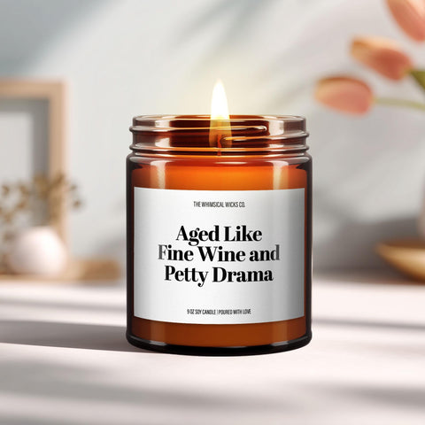 9oz amber jar candle with label “Aged Like Fine Wine and Petty Drama” lit on a table.