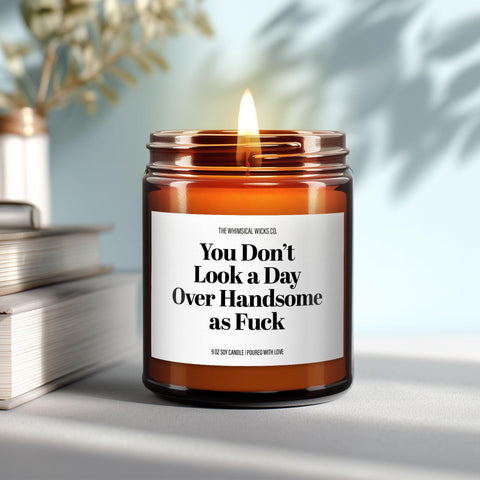 9oz amber jar candle with label “You Don’t Look a Day Over Handsome as Fuck” lit on a table.